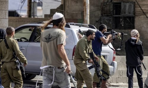 Israeli settlers fire at Palestinians (unseen) while an Israeli soldier (L) stands by during clashes in the town of Huwara in the occupied West Bank on October 13, 2022. (Photo by Oren ZIV / AFP)
